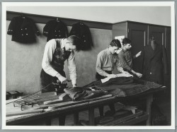 Leerlingen in de kleermakerij achter een tafel bezig met strijken en persen van kleding(-stoffen), ca. 1940